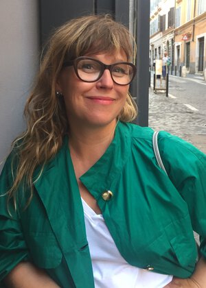 En smilende dame med briller, hvit topp og grønn jakke som lener seg mot en vegg med albuen. - Klikk for stort bilde